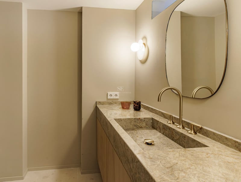 Nahaufnahme eines Waschbeckens aus Naturstein mit goldenem Wasserhahn, ovalem Spiegel und warmer Wandbeleuchtung.