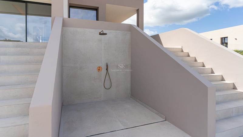 Minimalistische Außendusche mit hellgrauen Mikrozementwänden und Messingarmaturen vor blauem Himmel.