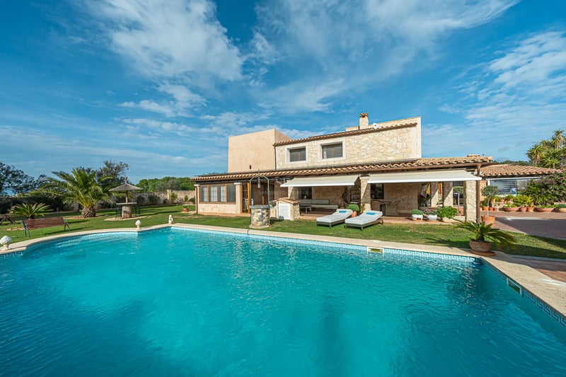Grande piscine turquoise devant une villa méditerranéenne traditionnelle en pierre avec pelouse et palmiers.