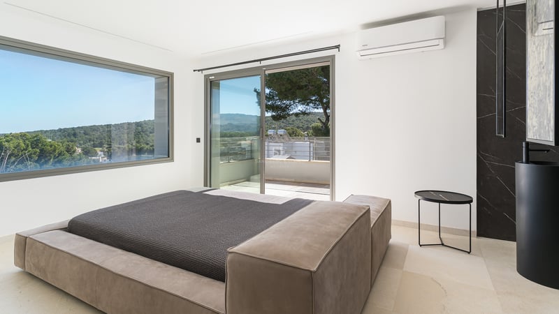 Modernt sovrum med låg sängram, stora skjutdörrar i glas mot terrass och fönster med utsikt över gröna kullar.