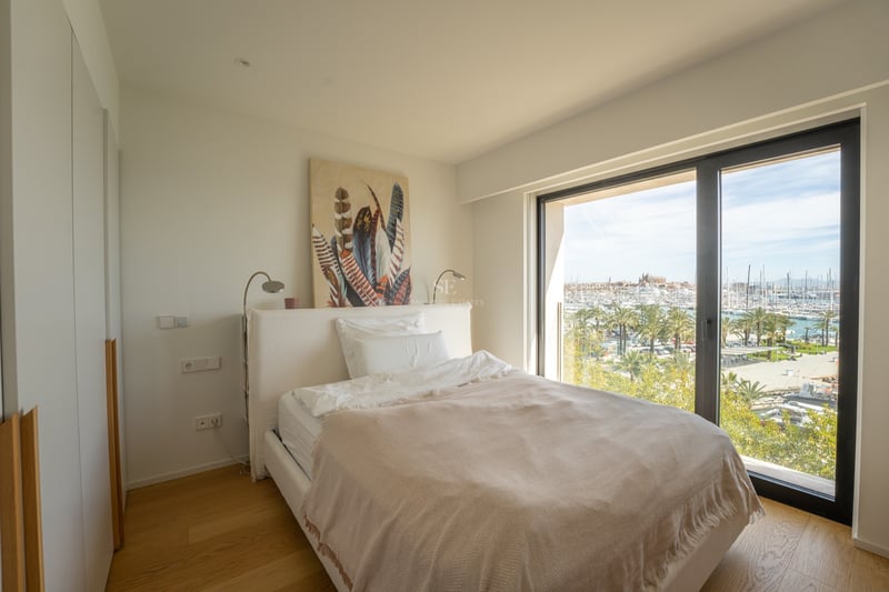 Modernt sovrum med vit säng och ett stort fönster med utsikt över en solig marina med palmer.