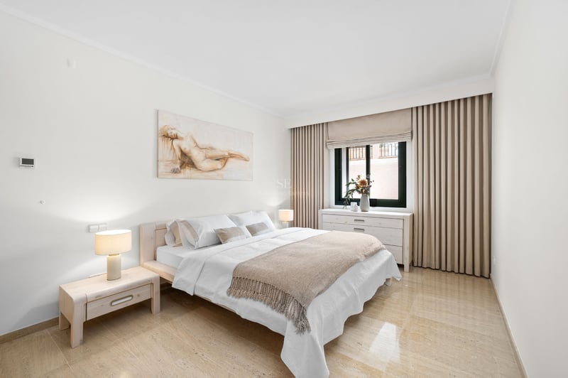 Chambre lumineuse avec lit en bois clair, sol en marbre, murs blancs et un grand tableau au-dessus du lit.