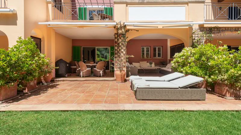 A sunny terracotta terrace with wicker lounge furniture and sunbeds bordering a lush green lawn.