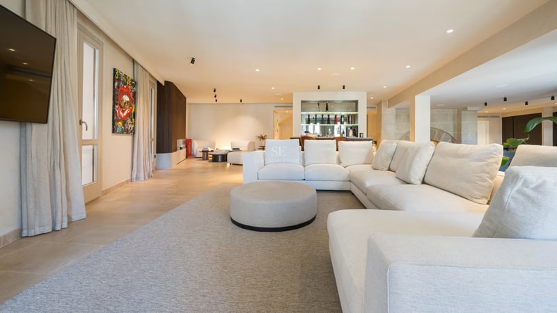 Espace de vie moderne et spacieux avec un grand canapé d'angle blanc, un tapis neutre et un bar élégant au fond.