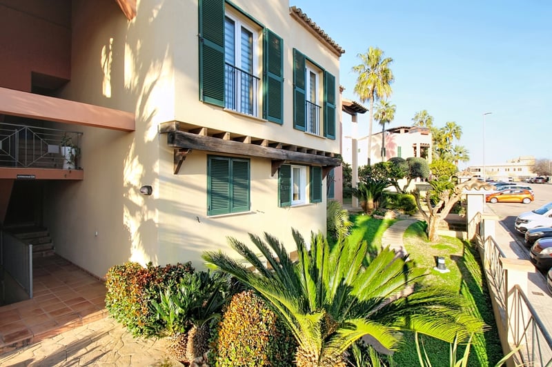 Exteriör av en beige byggnad med gröna fönsterluckor, träbjälkar och tropisk trädgård.