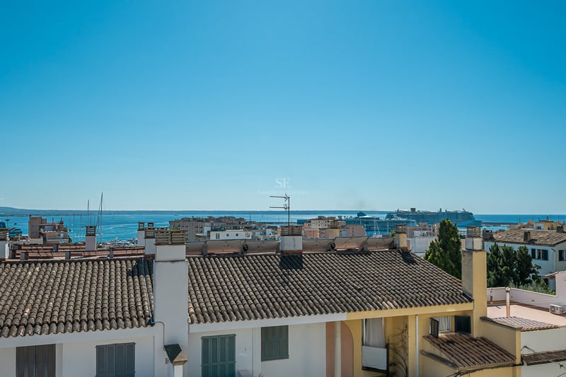 Elevated view over terracotta rooftops towards a blue sea with a large cruise ship in the harbor under a clear sky.