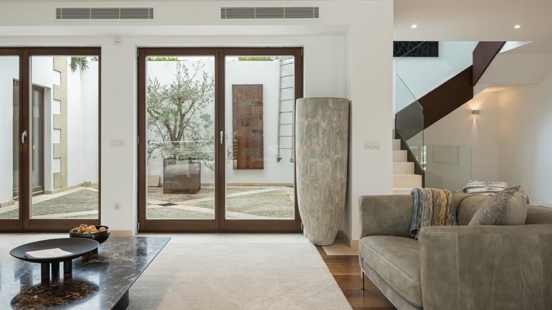 Salon moderne avec canapé en cuir, table en marbre et grandes baies vitrées ouvrant sur un patio avec un olivier.