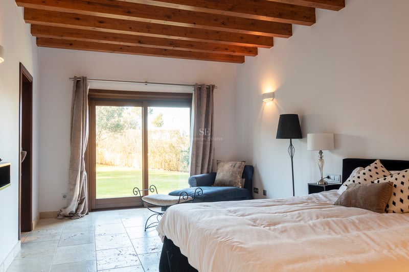 Sovrum med synliga takbjälkar i trä, naturstengolv och skjutdörr i glas mot trädgården.