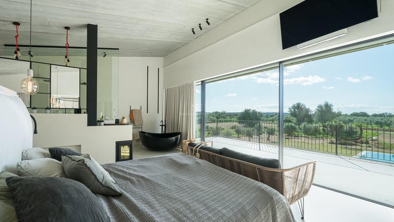 Modernt huvudsovrum med svart badkar, öppen spis och stora glaspartier mot det mallorkinska landskapet.