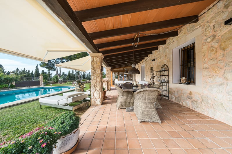 Grande terrasse couverte avec carrelage en terre cuite, murs en pierre et salle à manger en osier face à la piscine.