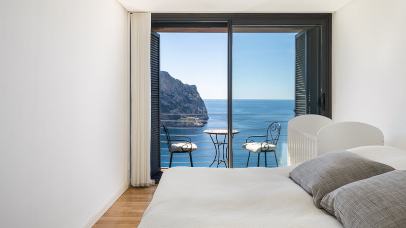 Minimalist white bedroom featuring a private balcony with wrought iron furniture and front-on views of a sea cliff.