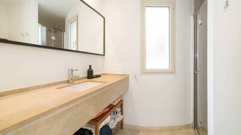 Modernt badrum med ett långt stentvättställ i beige, stor spegel och duschvägg i glas i ett ljust rum.