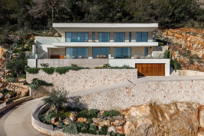 Modern white villa built into a rocky hillside with dry stone walls, large floor-to-ceiling windows, and a curved driveway.