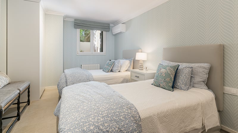 Bright room with two single beds, light blue patterned wallpaper, built-in wardrobes, and stone flooring.