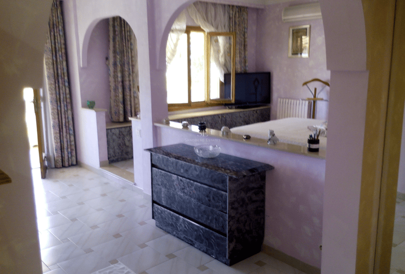 Dormitorio principal en tonos lavanda con arcos arquitectónicos, cómoda de mármol y suelos de baldosa clara.