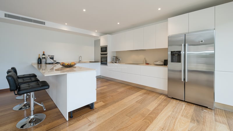 Cuisine blanche moderne avec parquet, réfrigérateur en inox, plan de travail clair et tabourets de bar noirs.