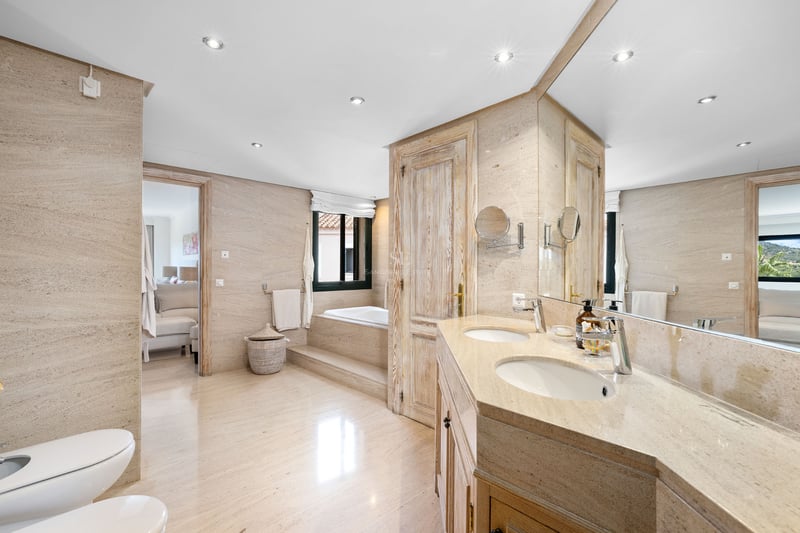 Salle de bain spacieuse avec murs en pierre beige, double vasque, grand miroir et baignoire encastrée au fond.