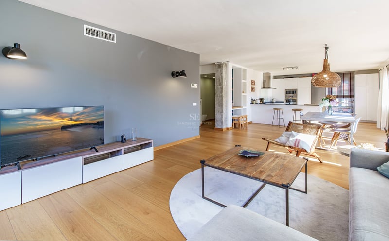 Ljust vardagsrum med öppen planlösning, ljusa trägolv, grå fondvägg och integrerat kök i bakgrunden.