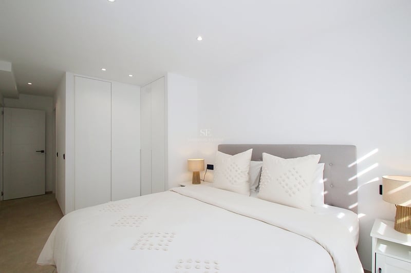 Ett ljust modernt sovrum med vita sängkläder, grå huvudgavel, integrerade garderober och naturligt ljus.