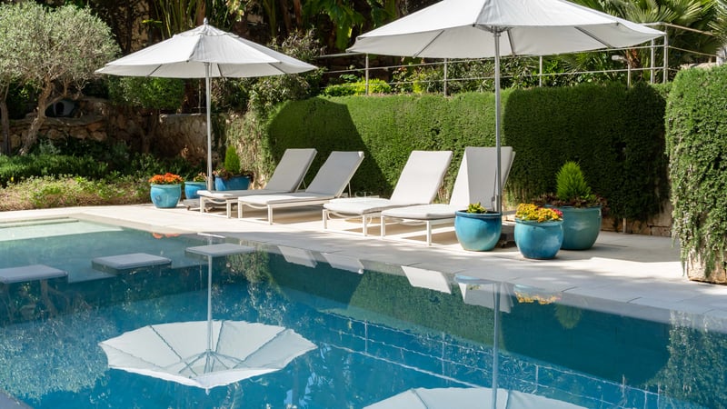 Luxuriöser Swimmingpool mit vier weißen Liegestühlen und Sonnenschirmen vor einer gepflegten grünen Hecke.