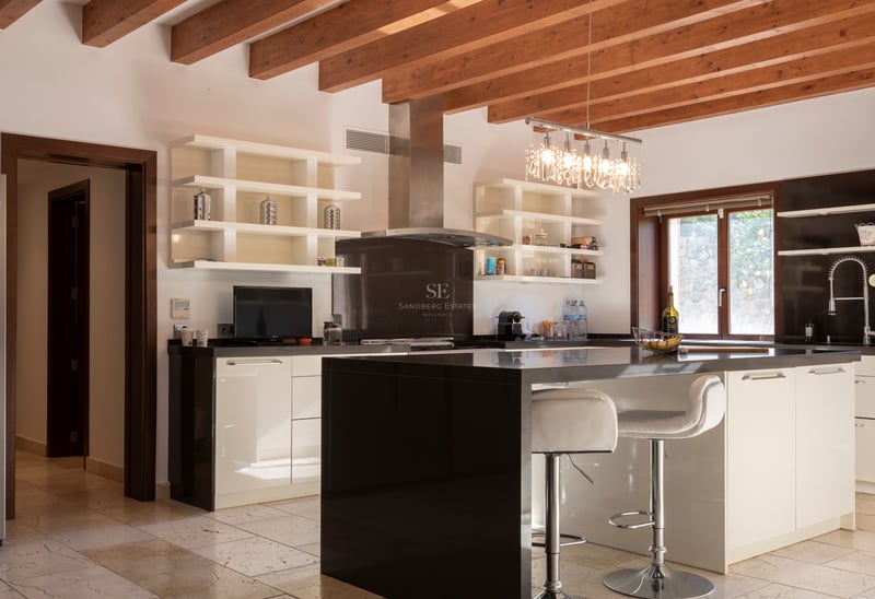 Modernt kök med svart köksö, vita skåp, synliga träbjälkar och naturstensgolv.