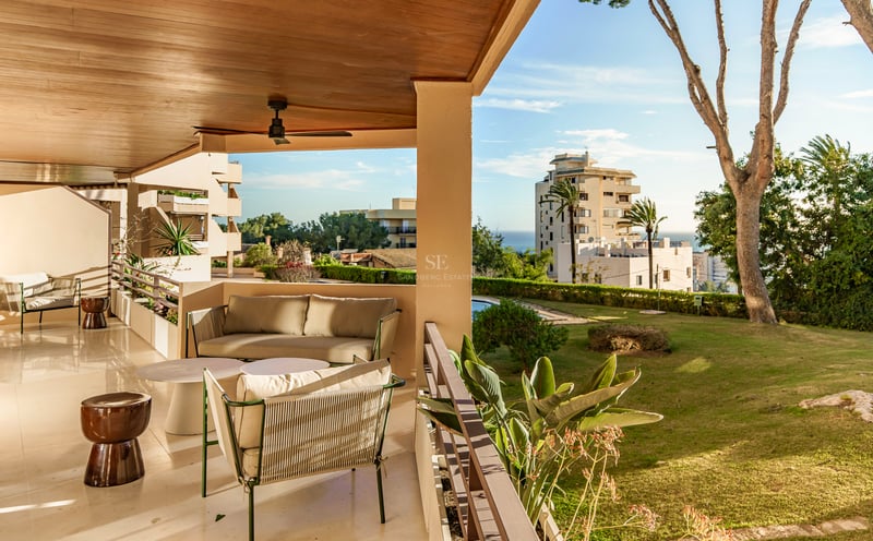 Überdachte Terrasse mit modernen Sofas, Holzdecke und Deckenventilator mit Blick auf einen Garten und das Meer.