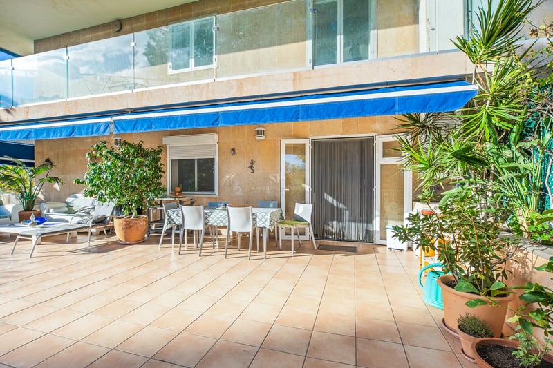 Sonnige Terrasse mit Keramikfliesen, Esstisch für sechs Personen, blauen Markisen und Glasgeländern.