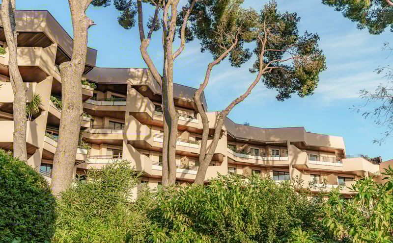 Mehrstöckiges modernes Gebäude mit eckigen Balkonen, umgeben von hohen Pinien unter klarem blauem Himmel.
