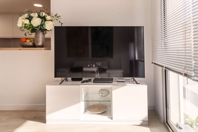 Flachbildfernseher auf weißem Lowboard neben sonnigem Fenster mit Jalousien und Blumendekoration.