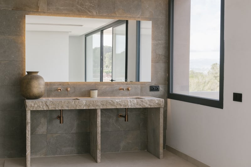 Modernes Badezimmer mit Doppelwaschbecken aus Rohstein, Wandarmaturen aus Bronze und hinterleuchtetem Spiegel.