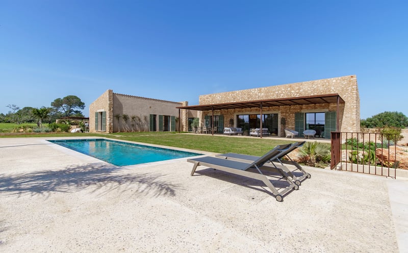 Türkisfarbener Pool mit grauen Liegen auf Steinterrasse vor einem Natursteinhaus und gepflegtem Rasen.