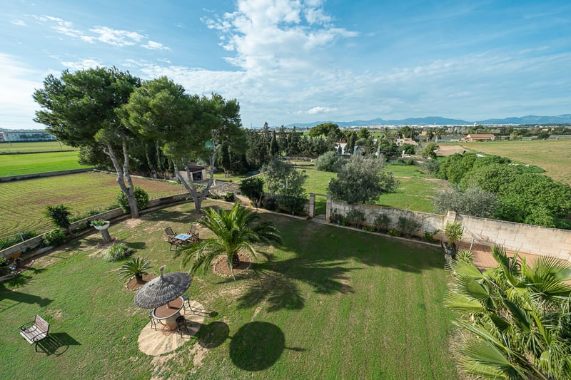Vue surélevée d'une grande pelouse avec palmiers, murs en pierre et parasol en chaume avec les montagnes en arrière-plan.