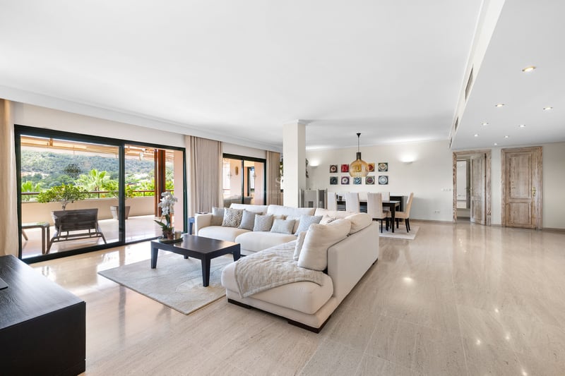 Salon spacieux avec canapé blanc en L, sol en marbre et grandes baies vitrées ouvrant sur une terrasse paysagée.
