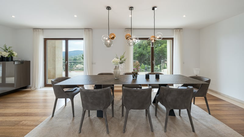 Salle à manger moderne avec table texturée, chaises grises et suspensions en verre devant de grandes baies vitrées.