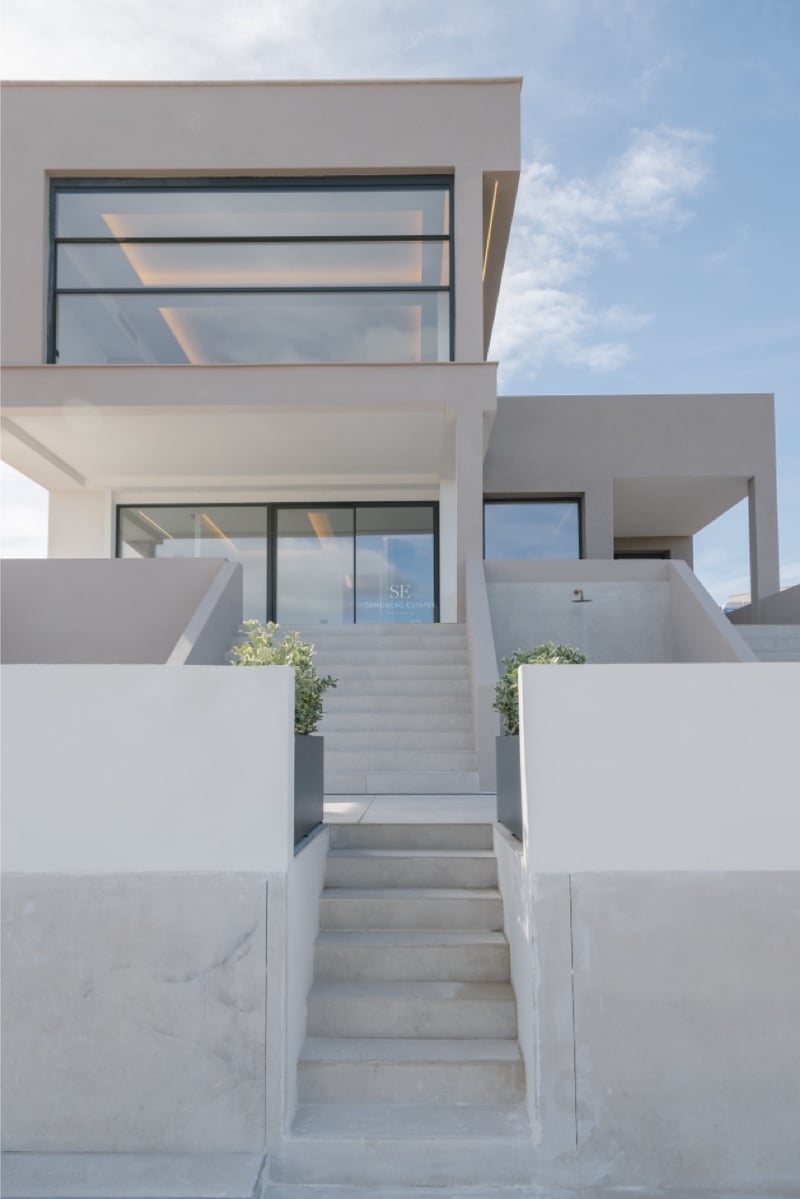 Vorderansicht einer modernen sandfarbenen Villa mit Steintreppen und großen Glasfenstern unter blauem Himmel.