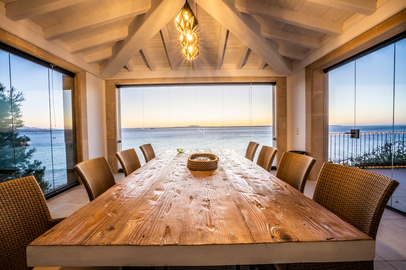 Großer Esstisch aus Holz auf einer verglasten Terrasse mit Panoramablick auf das Meer bei Sonnenuntergang.