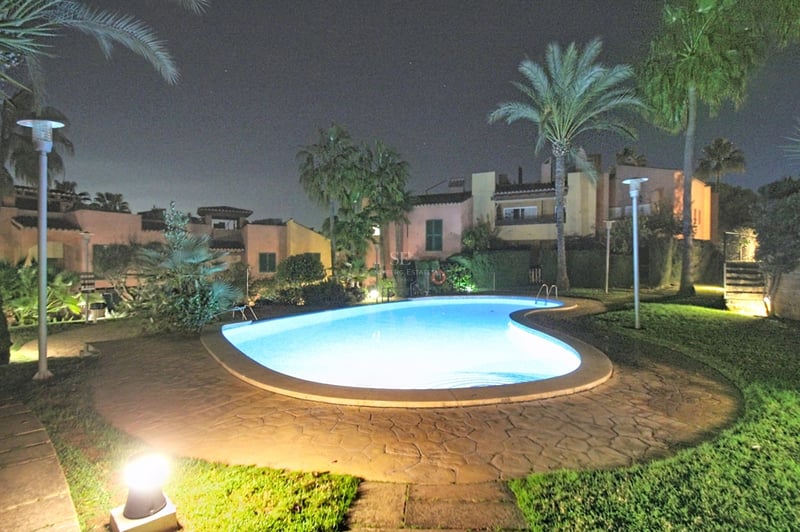 Njurformad pool upplyst på natten, omgiven av palmer, gräsmatta och medelhavsvillor under en mörk himmel.