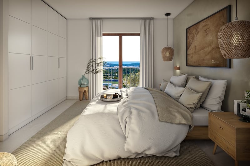 Bright master bedroom with a large bed, built-in white wardrobes, and a window overlooking a coastal landscape.