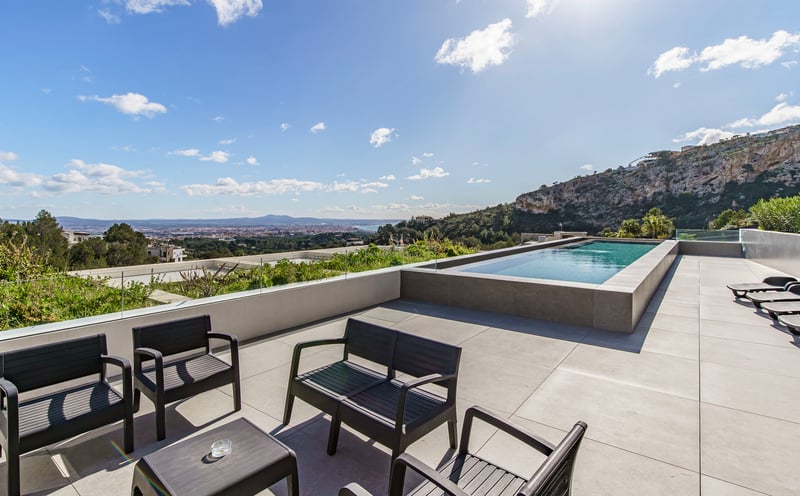 Terrasse de luxe avec piscine à débordement, garde-corps en verre, meubles noirs et vue panoramique.