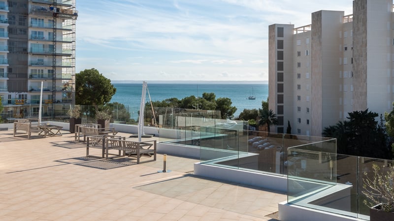 Grande terrasse carrelée avec garde-corps en verre et meubles en bois, surplombant la mer bleue et les bâtiments.