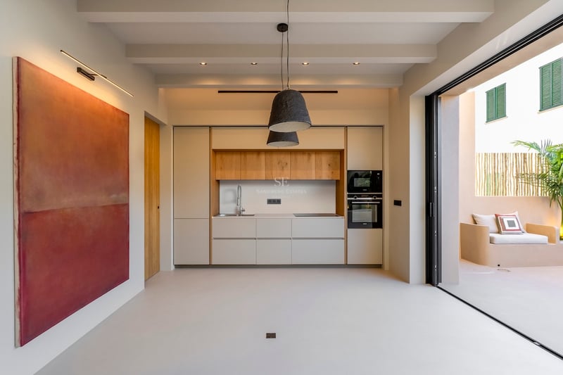 Cuisine minimaliste avec accents en bois clair, grand tableau rouge et baies vitrées ouvrant sur un patio ensoleillé.