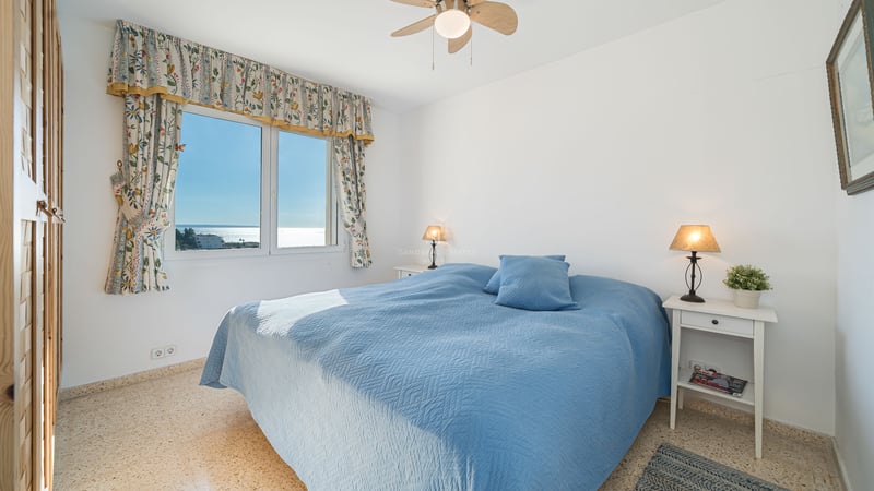A bright bedroom with a blue bedspread, sea views through a floral-curtained window, and bedside lamps.