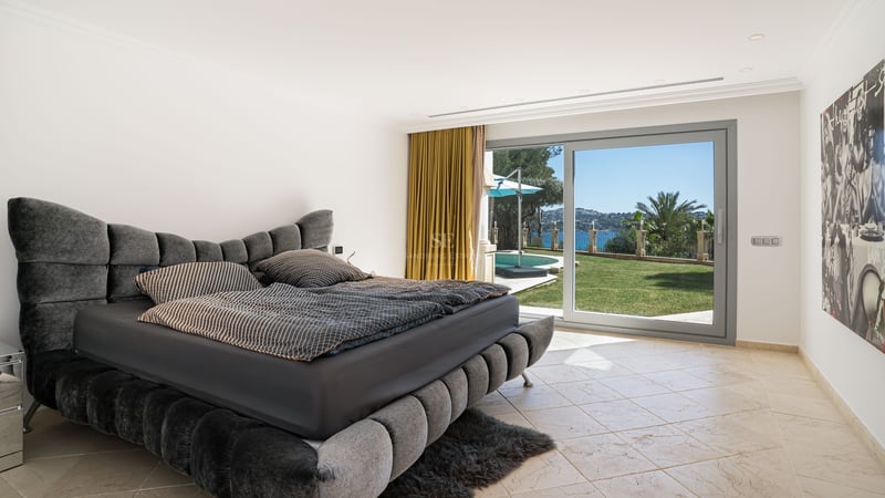 Modernes Schlafzimmer mit grauem Polsterbett und großen Glasschiebetüren, die zum Garten, Pool und Meer führen.