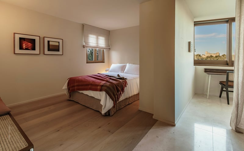 Minimalistisches Schlafzimmer mit weißem Bett, Holzböden und Arbeitsbereich mit Blick auf eine historische Burg.