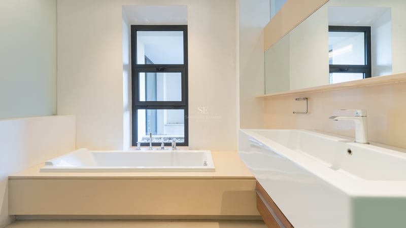 Modernt badrum med ett inbyggt vitt badkar intill ett fönster och ett stort rektangulärt tvättställ med stor spegel.