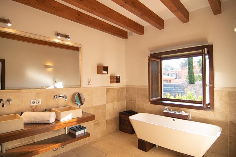 Badrum med synliga takbjälkar, fristående vitt badkar, dubbla stentvättställ och fönsterutsikt.