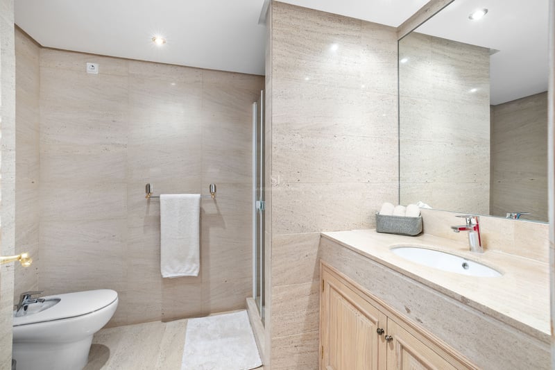 Salle de bain moderne avec murs en pierre beige, bidet blanc, meuble en bois avec lavabo et grand miroir.