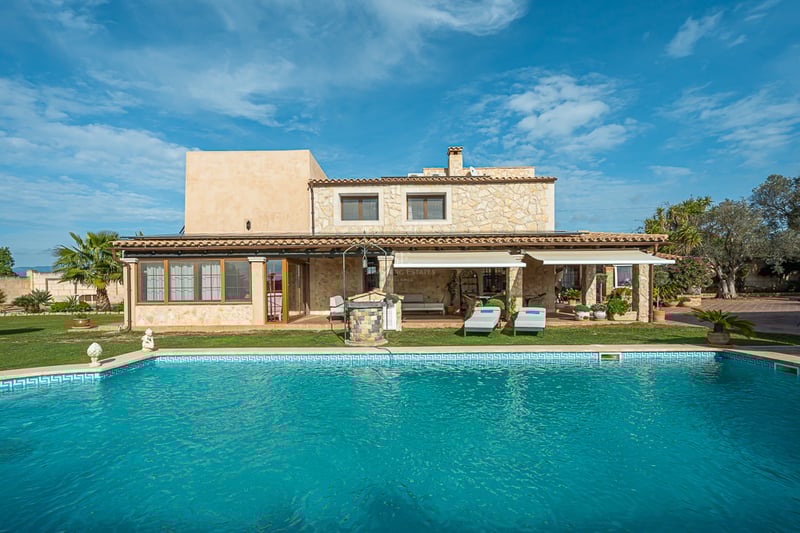 Grande piscine turquoise devant une villa méditerranéenne en pierre avec terrasse couverte et jardins.