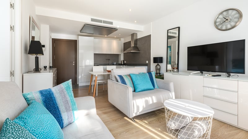 Salon lumineux avec fauteuil gris, coussins turquoise, meuble TV et cuisine moderne en arrière-plan.