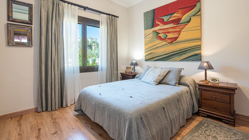 Ljust sovrum med trägolv, säng i gråa toner, klassiska nattduksbord och en stor färgstark tavla.